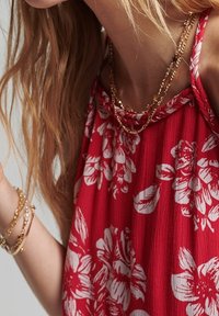 Röd blommig topp med vita blommönster, texturerad tyg och avslappnad passform. Guldhalsband med länkar och armband på handleden.