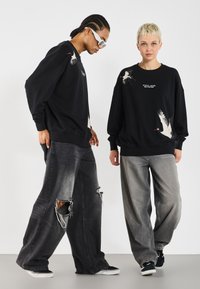 Black sweatshirts feature bird graphics and text, paired with distressed grey wide-leg jeans. One person wears sunglasses and black sneakers.