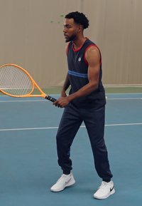 Modra športna majica brez rokavov z rdečimi detajli in horizontalno črto; mornarsko modri trenirke; bele športne čevlje; drži oranžno teniško lopar.