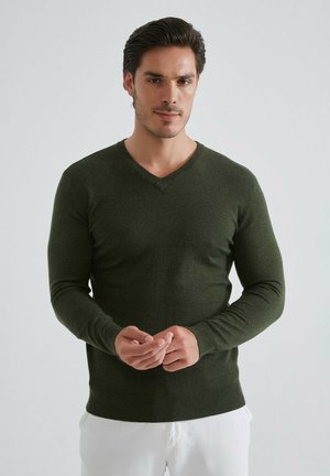 Pull en V vert foncé en tissu tricoté doux. Design ajusté avec des manches longues et un ourlet côtelé, porté sur un pantalon blanc.