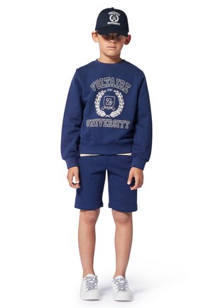 Marineblauwe sweatshirt en shorts set met de tekst "Voltaire University", ribgebreide manchetten en een bijpassende pet. Schoenen zijn wit met gebloemde veters.