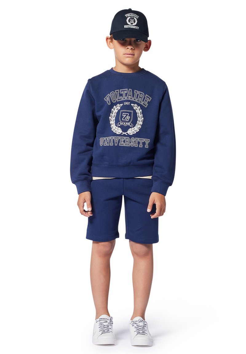 Completo felpa blu navy e pantaloni corti con testo "Università Voltaire", polsini a coste e cappellino abbinato. Le scarpe sono bianche con lacci a fantasia.