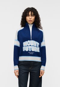 Μπλε πλεκτό πουλόβερ με ψηλό γιακά και κλείσιμο με φερμουάρ. Διαθέτει τη φράση "MOUNT FUTURE" σε λευκά και ανοιχτά μπλε γράμματα, καθώς και αντίθετες ρίγες.