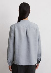 Ljusgrå blus med långa ärmar gjord av ett mjukt, texturerat tyg. Har en bakre söm och knappade manschetter. Rak fåll.