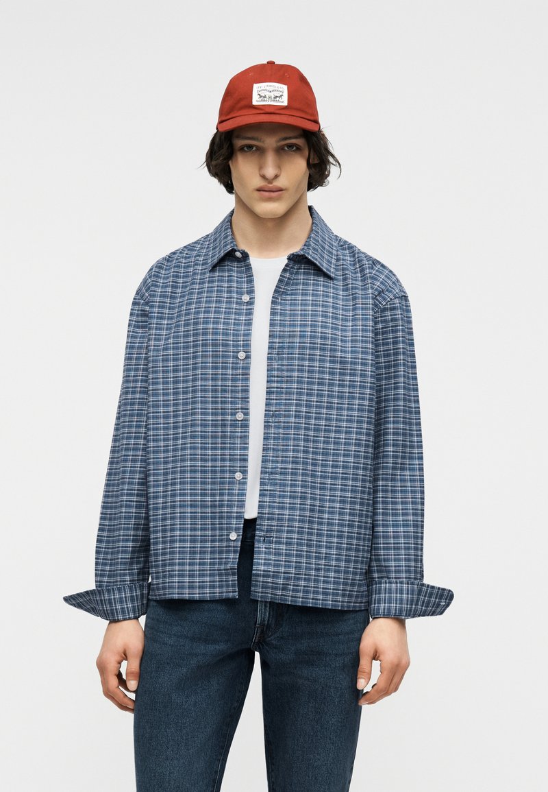 Jeune personne portant une casquette rouge, une chemise à carreaux bleue boutonnée sur un T-shirt blanc, et un jean bleu foncé, debout devant un fond uni.