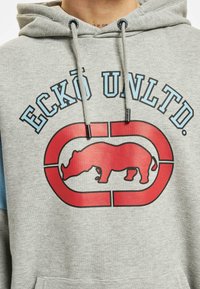 Šedá mikina s kapucí, na které je velký červený grafický motiv nosorožce uvnitř oválu, obklopeného modrým textem "ECKŌ UNLTD." Šňůrky kapuce jsou šedé.