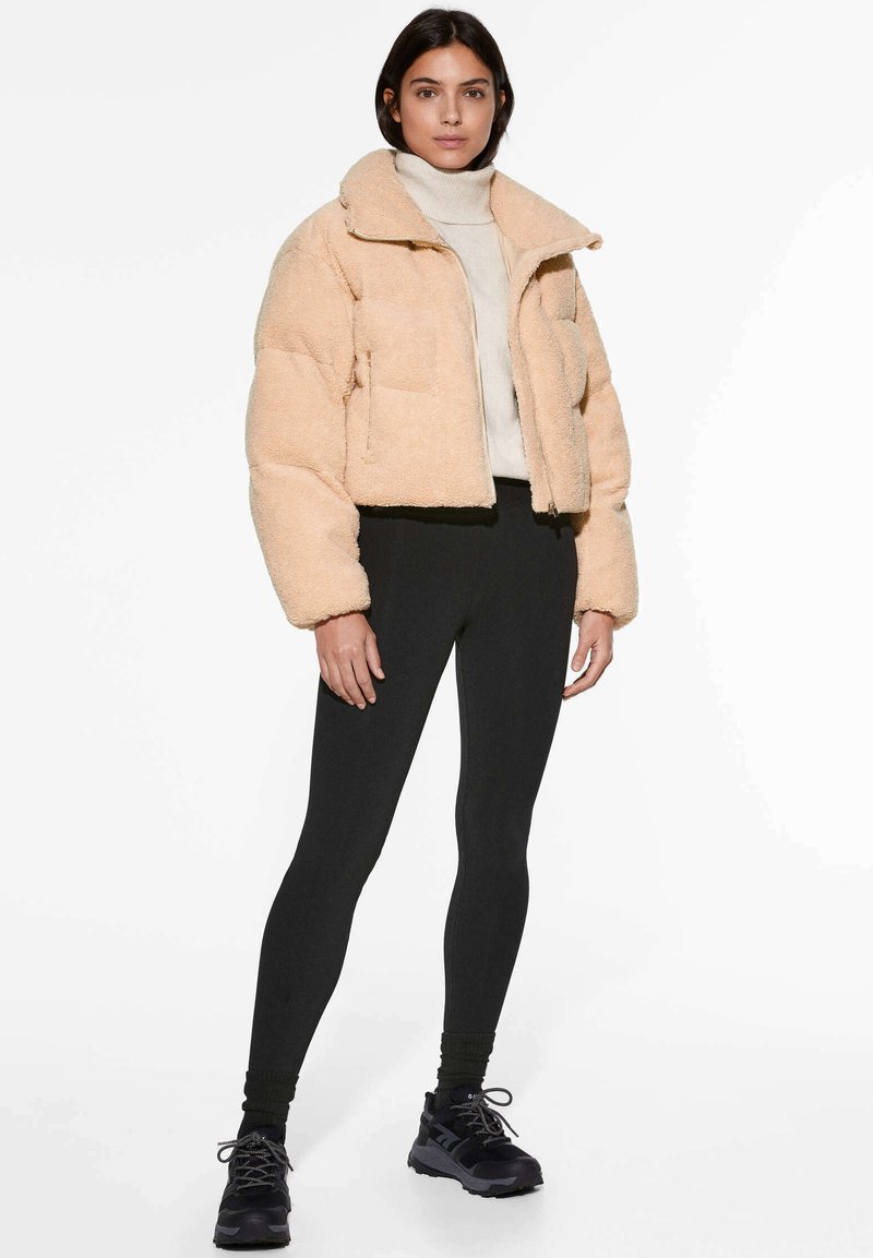 Gekürzte, flauschige beigen Jacke mit Kragen, Reißverschluss vorne und zwei Taschen, kombiniert mit schwarzen Leggings und dunklen Sportschuhen.