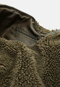 Olijfgroene fleece stof met een zachte, gestructureerde oppervlakte; heeft een donkere, glanzende ritssluiting en een satijnen binnenvoering.