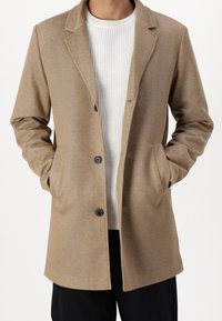 Beige uldblandingsfrakke med klassisk krave, enkelt knaplukning, sidelommer og lige snit, lagdelt over en hvid ribbet sweater.