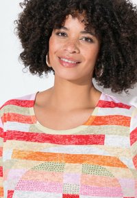 Värikäs raidallinen paita, jossa on punaisia, oransseja, vaaleanpunaisia ja kermansävyisiä kuvioita, tekstuurisia yksityiskohtia ja pieniä vihreitä koristeita. Pyöreä pääntie.