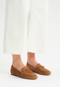 Brown suede loafers featuring metal studs and a decorative strap. The shoes have a flat sole and a rounded toe design.