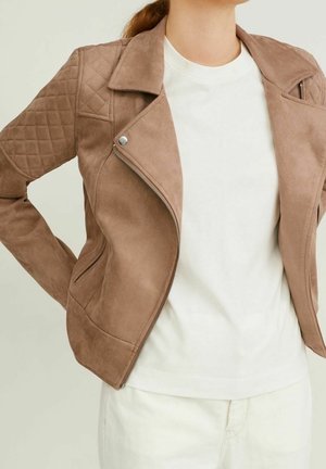 Kvinde iført en lysebrun ruskinds-motocykeljakke med quiltede skuldre over en enkel hvid t-shirt og cremefarvede bukser, hænderne i siden.