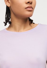 Κοντινό πλάνο ενός ατόμου με πλεγμένα μαλλιά που φοράει ένα ανοιχτό μωβ μπλουζάκι με στρογγυλή λαιμόκοψη, εμφανίζοντας το λαιμό, το πηγούνι και το επάνω μέρος του στήθους.