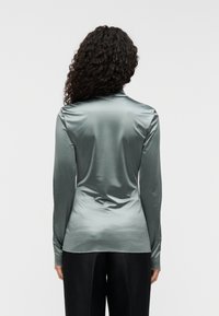 Μπλούζα με μακριά μανίκια από απαλό, γυαλιστερό ύφασμα σε χρώμα πετρόλ. Διαθέτει εφαρμοστή γραμμή με ψηλό γιακά και καμπυλωτή ραφή στο τελείωμα, συνδυασμένη με μαύρο παντελόνι.