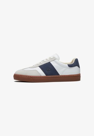 Lage sneaker met wit leer, beige suède accenten, een donkerblauwe zijstreep en een bruine rubberen zool, gezien vanaf de zijkant.
