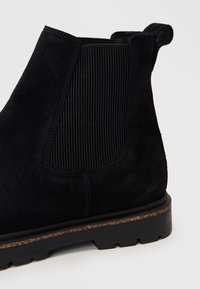 Stivale Chelsea in suede nero con pannello laterale elastico a coste, linguetta per il tallone e suola robusta con battistrada e bordo marrone.