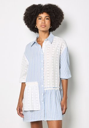 Donna con capelli ricci naturali che indossa un vestito con bottoni blu e bianco, realizzato con tessuti a righe e motivi misti, in piedi contro un muro semplice.