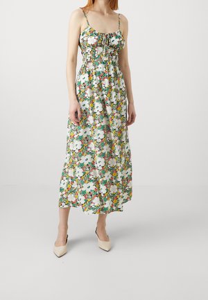 Femme portant une robe midi sans manches avec de fines bretelles et un motif floral coloré, associée à des talons beiges à bout pointu.