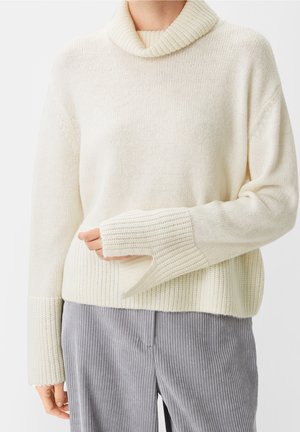Personne portant un pull à col roulé crème avec des poignets côtelés allongés et un pantalon gris à rayures verticales, debout devant un fond blanc.