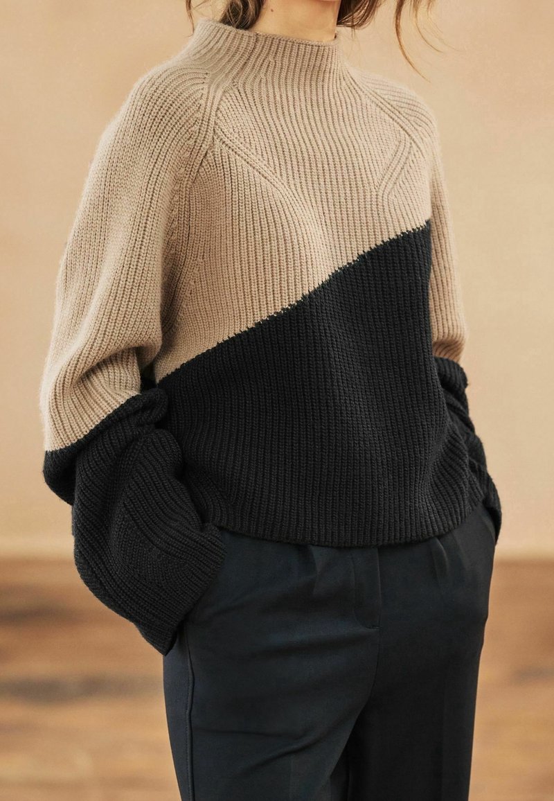 Femme portant un pull en maille beige et noir à blocs de couleurs avec de longues manches bouffantes et un pantalon noir, mains dans les poches.