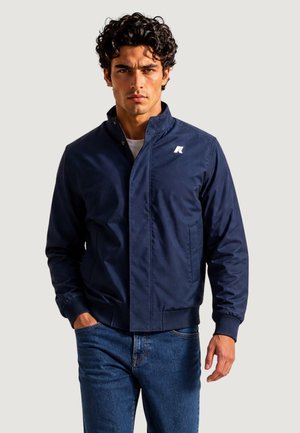 Mand med krøllet mørkt hår iført en mørkeblå jakke over en hvid skjorte og blå jeans, står mod en ensfarvet lys baggrund.