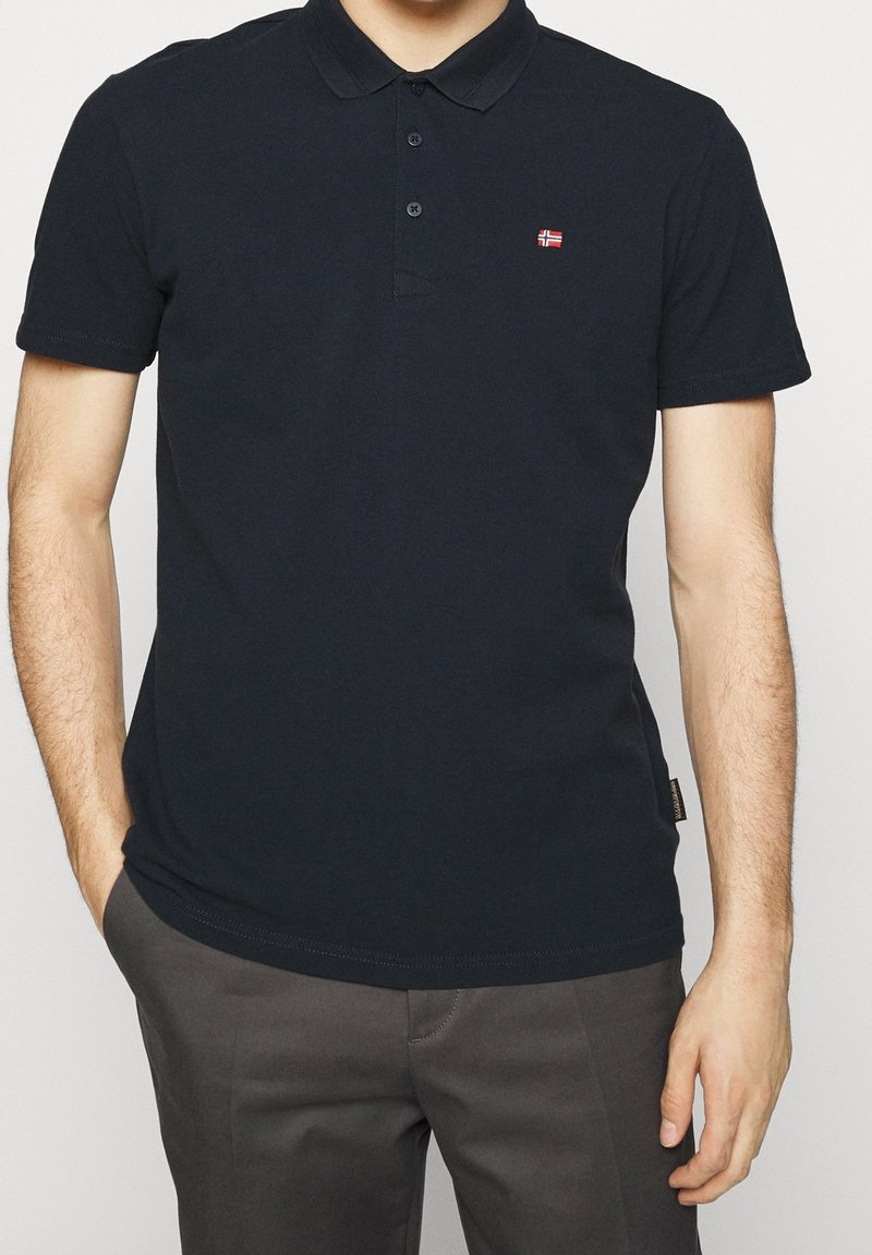 Homme portant un polo noir à manches courtes avec un emblème du drapeau norvégien, debout avec une main dans la poche d'un pantalon gris sur fond blanc.