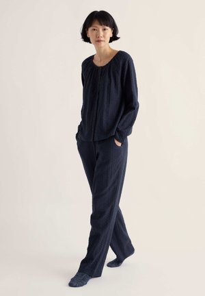 Femme aux cheveux courts et noirs portant un haut texturé à manches longues sombre et un pantalon assorti, debout les mains dans les poches sur un fond clair.