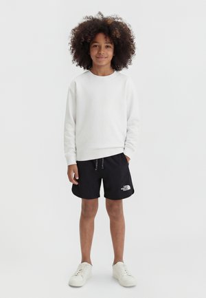 Lokkis juustega laps, kes kannab valget pusa, musti The North Face lühikesi pükse ja valgeid tossusid, seisab ühe käe taskus.