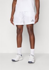 Biele športové šortky s elastickým pásom, s čiernym logom "Sergio Tacchini" blízko lemu. Doplnené o biele ponožky a tenisky.