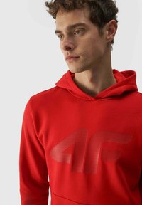Sudadera con capucha roja que presenta un gran logo texturizado gris en la parte delantera. La tela parece suave, con un diseño clásico y bolsillo canguro.