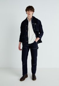 Mörk denimjacka med kontrasterande sömmar, med två framfickor. Parat med matchande mörka denimbyxor och en vit polo t-shirt under.
