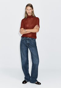 Rostfarbenes, kurzärmeliges Oberteil kombiniert mit lockeren, blauen Jeans. Das Model trägt schwarze Sandalen und hat glattes, langes Haar. Schlichter Hintergrund.