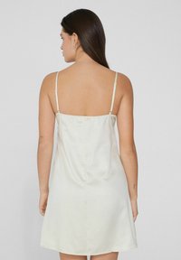 Vestido de satén en color crema claro, con finas tirantes ajustables y un corte holgado. Textura suave con un brillo sutil, de longitud media hasta el muslo.