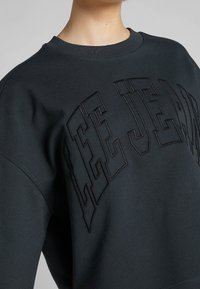 Sudadera gris oscuro con texto en relieve "LEE JEANS", cuello redondo acanalado y ajuste oversized con textura de tela suave.