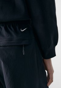 Črne športne hlače z vrvico v pasu, s srebrnim logotipom Nike na zadnjem žepu in teksturirano tkanino.