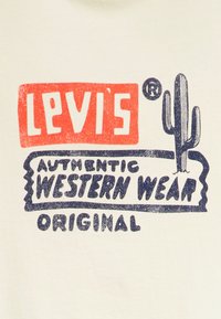 Kreemivärviline kangas, millel on graafiline kujundus, kus on paksus punases kirjas "Levi's" ja tumesinises kirjas "AUTHENTIC WESTERN WEAR ORIGINAL", koos kaktuse illustreerimisega.