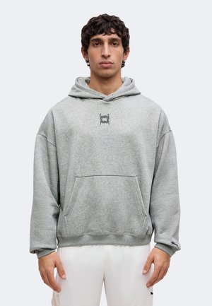 Jeune homme portant un sweat à capuche gris avec poche avant et petit motif noir près du cou, associé à un pantalon blanc.