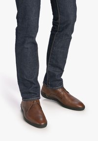 Zapatos de cuero marrón con superficie texturizada, de perfil bajo, punta redonda y cordones, combinados con jeans de mezclilla oscura que presentan una costura en contraste.