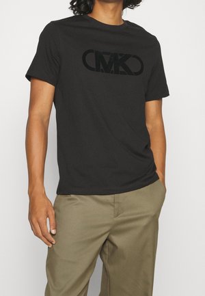 T-shirt en coton noir à manches courtes et col rond, avec un logo texturé noir en relief sur le devant. Porté avec un pantalon kaki clair.