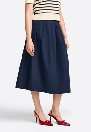 Gonna A-line navy con pieghe, realizzata in tessuto leggero. Abbinata a décolleté rosse con punta a forbice e dettaglio a fiocco.