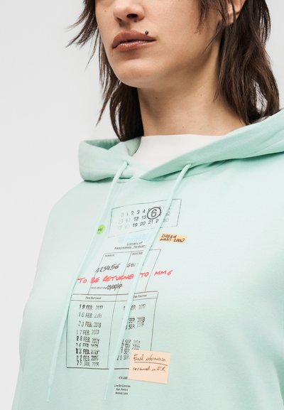 Άτομο που φοράει φούτερ με κουκούλα σε μέντα πράσινο χρώμα, με εκτυπωμένη κάρτα βιβλιοθήκης και χειρόγραφες σημειώσεις σε κόκκινο και πορτοκαλί χρώμα στο μπροστινό μέρος.