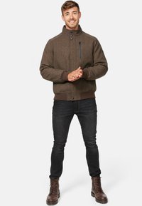 Braune strukturierte Jacke mit hohem Kragen, Frontknöpfen und einer Reißverschlusstasche auf der Brust, kombiniert mit dunklen Jeans und braunen Schnürstiefeln.