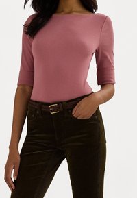 Nainen, jolla on päällään istuva vaaleanliila puolihihainen paita, joka on tungettu tummanruskeisiin samettihousuihin. Housuissa on kudottu ruskea vyö ja toinen käsi on taskussa.