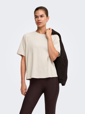 Mujer con una camisa beige de manga corta y suelta y leggings oscuros, sosteniendo una chaqueta negra texturizada sobre su hombro contra un fondo sencillo.