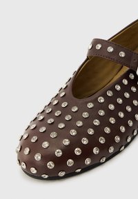 Brown leather shoe with a rounded toe, featuring a strap and adorned with silver studs across the upper surface. Smooth texture.