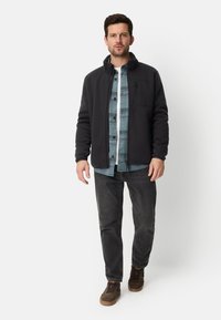 Zwarte fleecejas met een hoge kraag over een blauw geruit overhemd. Donkere spijkerbroek en bruine sneakers. Eenvoudig ontwerp met een ritssluiting zak.