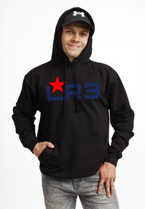 Ung mand iført sort hættetrøje med rød stjerne og blå "LR3" tekst, sort Under Armour kasket og grå jeans, smilende med hånden i lommen.