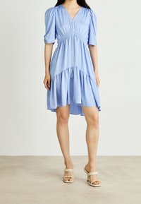 Vaaleansininen, polvipituinen mekko, jossa on lyhyet puffihihat, gathered vyötärö ja frillalla varustettu helma. Liukas kangas, V-mallinen kaula-aukko. Yhdistetty vaaleanbeigeihin korkokenkiin.