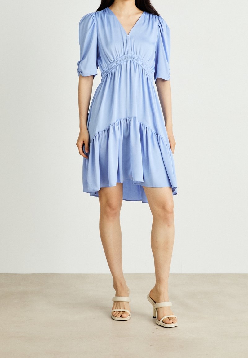 Vaaleansininen, polvipituinen mekko, jossa on lyhyet puffihihat, gathered vyötärö ja frillalla varustettu helma. Liukas kangas, V-mallinen kaula-aukko. Yhdistetty vaaleanbeigeihin korkokenkiin.