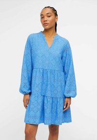 Blå blondekjole med V-hals, lange ermer og lagdelt design. Har intrikate broderie anglaise-detaljer gjennom hele plagget.